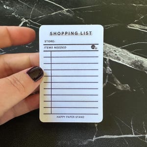 May include: A white rectangular shopping list card with rounded corners featuring black printed text and lines. The card includes the headers SHOPPING LIST, STORE, and ITEMS NEEDED with a small smiley face icon, and the text HAPPY PAPER STAND at the bottom.