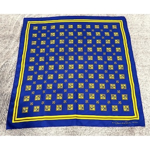 May include: A square blue scarf featuring a repeating pattern of yellow squares containing black abstract symbols, interspersed with small gold fleur-de-lis icons. The border consists of yellow and gold stripes. The text Oscar de la Renta is visible.