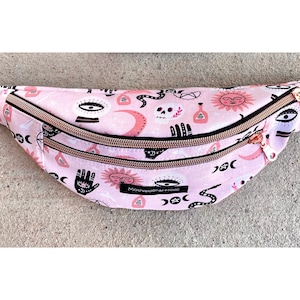 Peut inclure: Sac banane rose avec un motif répété de symboles célestes et occultes, dont des yeux, des lunes, des soleils et des serpents. Le sac a une sangle noire réglable, des fermetures éclair dorées et le texte "Motivational-Holic".