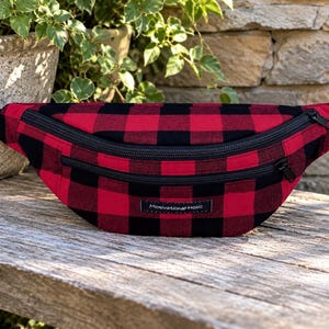 Könnte beinhalten: Eine Bauchtasche mit einem rot-schwarzen Karomuster. Die Tasche verfügt über zwei horizontale Reißverschlussfächer mit schwarzen Reißverschlüssen und einen verstellbaren schwarzen Gurt. Ein kleines rechteckiges Etikett mit dem Text Motivational Holic ist mittig angebracht.