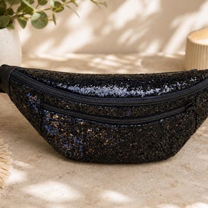 May include: A black glittery fanny pack featuring a crescent-shaped body with a textured, sparkling surface. The bag includes two horizontal zippered compartments with black hardware and an adjustable black nylon strap with a side-release buckle.