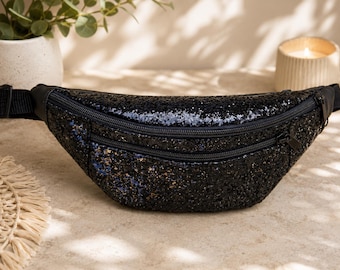 May include: A black glittery fanny pack featuring a crescent-shaped body with a textured, sparkling surface. The bag includes two horizontal zippered compartments with black hardware and an adjustable black nylon strap with a side-release buckle.