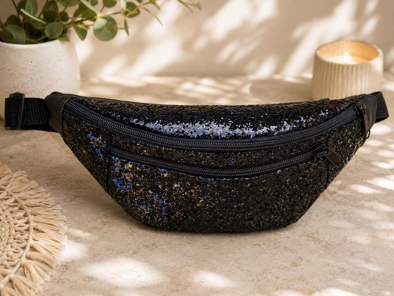 May include: A black glittery fanny pack featuring a crescent-shaped body with a textured, sparkling surface. The bag includes two horizontal zippered compartments with black hardware and an adjustable black nylon strap with a side-release buckle.