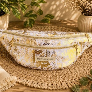 May include: A fanny pack featuring a white and metallic gold snakeskin pattern. The bag has two horizontal zippered compartments with gold-tone hardware and a white adjustable strap. A rectangular label on the front displays the text Motivational-Holic.