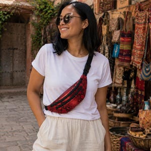 May include: A red and black snakeskin patterned fanny pack with a black zipper and an adjustable black strap. The bag is worn across the chest over a plain white t-shirt and light-colored trousers, featuring a metallic finish on the textured material.