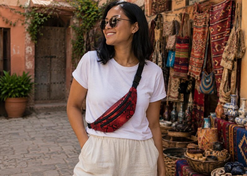 May include: A red and black snakeskin patterned fanny pack with a black zipper and an adjustable black strap. The bag is worn across the chest over a plain white t-shirt and light-colored trousers, featuring a metallic finish on the textured material.