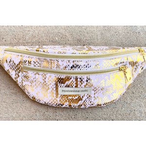 May include: A light pink fanny pack with a gold snakeskin pattern. The bag has two zippered compartments with gold zippers and a white adjustable strap. The text "Motivational-Holic" is on a small label on the front.