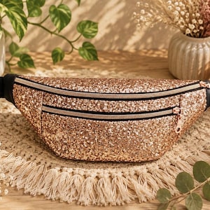 May include: A rose gold glitter fanny pack featuring two horizontal zippered compartments with black trim and a black adjustable nylon strap. The bag has a textured, sparkling surface and metallic rose gold zipper pulls.