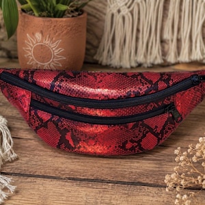 Peut inclure: Un sac banane rouge et noir avec un motif texturé en peau de serpent. Le sac possède deux compartiments horizontaux zippés avec des fermetures éclair noires et une sangle noire réglable fixée des deux côtés pour une fixation sécurisée.