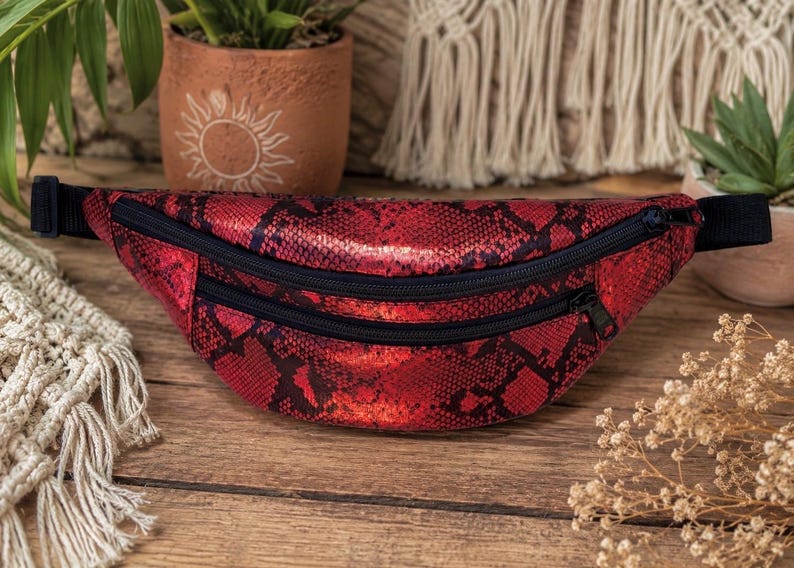 May include: A red and black fanny pack featuring a snakeskin texture pattern. The bag has two horizontal zippered compartments with black zippers and a black adjustable strap attached to both sides for secure fastening around the waist or chest.