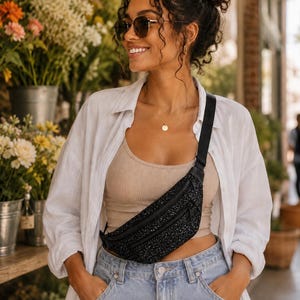 May include: A black fanny pack with a textured, glittery surface and two zippered compartments. The bag features a black adjustable strap worn across the chest, providing a secure and functional way to carry small personal items while on the go.