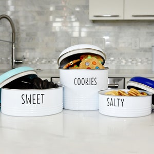 May include: Four white metal canisters with lids, labeled "Cookies", "Salty", "Sweet", and one with a blue lid. The canister labeled "Cookies" is open and filled with chocolate chip cookies.