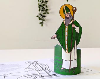 May include: A paper craft of Saint Patrick, a religious figure, standing on a green background with shamrocks. He is wearing a green robe with a white collar and a gold halo. He is holding a staff with a gold crook.