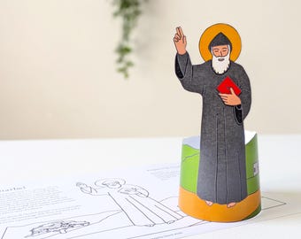 May include: A paper cutout of Saint Charbel, depicted in a gray robe with a white beard and a yellow halo. He holds a red book and raises his right hand. The base is a green and yellow cylinder. A coloring page is in the foreground.