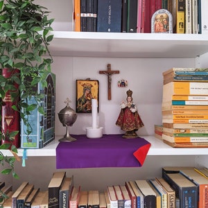 May include: A bookshelf with a variety of books, including "The Glimpse of Truth", "The Secret History", and "Breakfast of Titans". There is a purple cloth on the shelf with a gold metal incense burner and a white candle. There is a religious icon with a woman and child, a crucifix, and a figurine of a person in a red and gold robe.