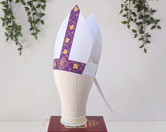 May include: A white and purple paper bishop's hat with gold accents and a design of grape leaves. The hat is on a white mannequin head and is sitting on top of a stack of books.