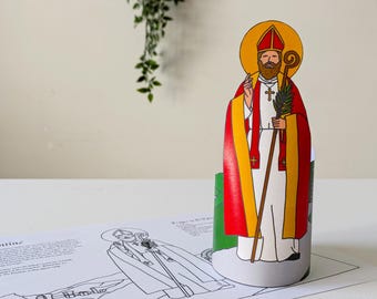 May include: A colorful, illustrated figure of a saint, wearing a red and yellow robe, holding a staff and a sprig of leaves. The figure stands on a cylindrical base, with a halo behind the head. A coloring page is in the foreground.