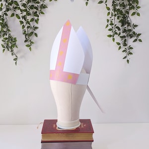 May include: A white and pink paper hat with a gold pattern. The hat is on a white mannequin head.