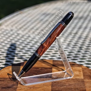 May include: A wooden pen with a black and brown wood grain pattern and silver accents. The pen is on a clear acrylic stand.