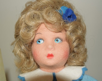 May include: A doll with blonde curly hair wearing a blue dress with a white collar and a blue flower in her hair.