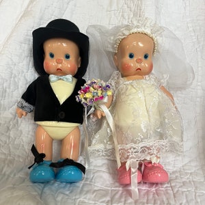 May include: A bride and groom doll set. The bride doll is wearing a white lace dress with a veil and pink shoes. The groom doll is wearing a black tuxedo with a white bow tie and blue shoes. They are holding a bouquet of flowers.