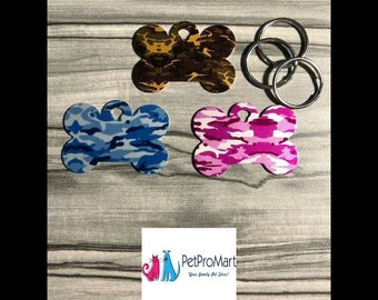 May include: Three bone-shaped dog tags with different camouflage patterns: brown and black, blue and white, and pink and white. The tags have a silver ring attached to the top.