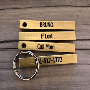 May include: A set of four gold-colored metal tags with engraved text. The tags are attached to a silver key ring. The text on the tags reads: "BRUNO", "If Lost", "Call Mom", and "501-517-1773".