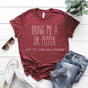 May include: Burgundy t-shirt with the text "BRING ME A DR PEPPER and I'll love you forever" in white. The shirt is paired with blue jeans, white sneakers, and sunglasses. A small plant is in the background.