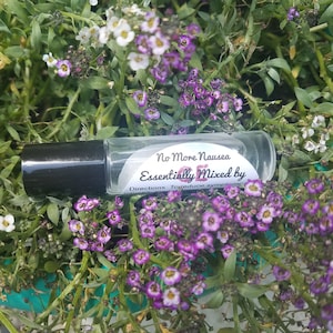 May include: A clear glass roller bottle with a black cap, labelled "No More Nausea, Essentially Mixed by". The bottle is surrounded by green foliage and clusters of small purple and white flowers. The label includes directions to reduce symptoms.