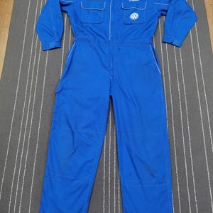 May include: A blue Volkswagen mechanic's jumpsuit with long sleeves, a collar, and multiple pockets. The jumpsuit features the Volkswagen logo and the name "HYOSHOKA" embroidered on the chest. White piping accents the seams.