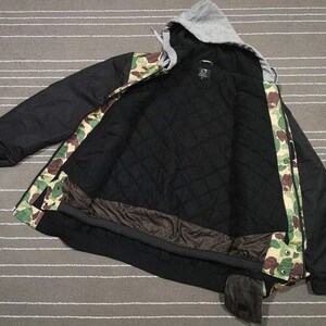 K&ouml;nnte beinhalten: Schwarze und gr&uuml;n getarnte Kapuzenjacke mit schwarzem Rei&szlig;verschluss und schwarzem Futter. Die Jacke hat eine Kapuze mit Kordelzug und eine Rei&szlig;verschlusstasche am linken &Auml;rmel.
