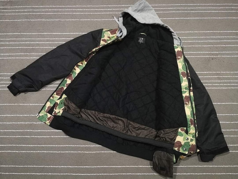 K&ouml;nnte beinhalten: Schwarze und gr&uuml;n getarnte Kapuzenjacke mit schwarzem Rei&szlig;verschluss und schwarzem Futter. Die Jacke hat eine Kapuze mit Kordelzug und eine Rei&szlig;verschlusstasche am linken &Auml;rmel.