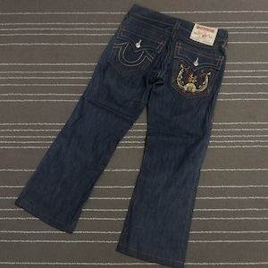 Könnte beinhalten: Dunkelblaue Jeans aus Denim mit roten Nähten und einem großen gestickten Design auf der Gesäßtasche. Das Design zeigt einen Totenkopf mit Flammen und einem Hufeisen.