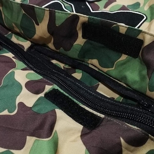 K&ouml;nnte beinhalten: Nahaufnahme eines Tarnstoffes mit einem schwarzen Rei&szlig;verschluss und einem schwarzen Klettverschluss. Der Stoff ist eine Mischung aus Braun, Gr&uuml;n und Beige.
