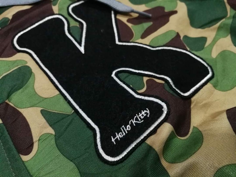 K&ouml;nnte beinhalten: Ein gestickter schwarzer und wei&szlig;er Aufn&auml;her mit einem gro&szlig;en Buchstaben K und dem Text "Hello Kitty" auf einem Tarnstoff-Hintergrund.
