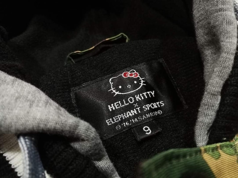 K&ouml;nnte beinhalten: Schwarz-gr&uuml;ner Kapuzenpullover mit einem Hello Kitty x Elephant Sports-Etikett. Das Etikett zeigt eine Hello Kitty-Karikatur mit einer roten Schleife und den Text "Hello Kitty x Elephant Sports &copy;'16 '11 Sanrio 9".
