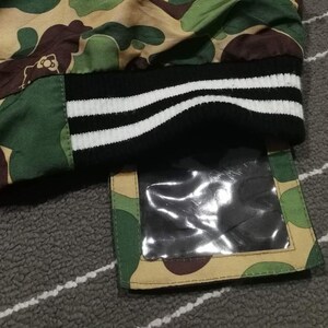 K&ouml;nnte beinhalten: Eine gr&uuml;ne, braune und beigefarbene Tarnjacke mit einem schwarz-wei&szlig; gestreiften B&uuml;ndchen. Die Jacke hat eine transparente Plastiktasche auf der rechten unteren Seite.
