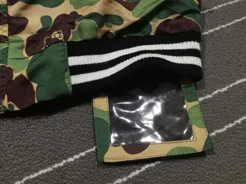 K&ouml;nnte beinhalten: Eine gr&uuml;ne, braune und beigefarbene Tarnjacke mit einem schwarz-wei&szlig; gestreiften B&uuml;ndchen. Die Jacke hat eine transparente Plastiktasche auf der rechten unteren Seite.