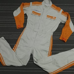 May include: A gray jumpsuit with orange accents and the text "Toyota" on the chest. The jumpsuit has two pockets on the chest and two side pockets. The jumpsuit has orange stripes down the legs and arms.