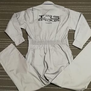 May include: A light gray work jumpsuit with long sleeves and a zipper closure. The jumpsuit has a black elastic waistband and two large pockets on the front. The back of the jumpsuit has a black logo that says "TRS GROUP TOYOTA".