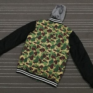 K&ouml;nnte beinhalten: Eine schwarze und gr&uuml;ne Bomberjacke mit Tarnmuster und einem schwarz-wei&szlig;en Streifenbesatz an Kragen und &Auml;rmelb&uuml;ndchen. Die Jacke hat ein graues Kapuzenfutter.