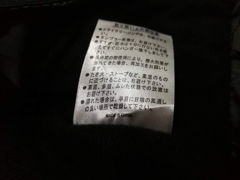 K&ouml;nnte beinhalten: Ein wei&szlig;es Pflegeetikett mit schwarzem Text in Japanisch, das Anweisungen zur Reinigung und Pflege des Kleidungsst&uuml;cks gibt. Der Text enth&auml;lt Informationen &uuml;ber chemische Reinigung, Trocknergeeignetheit, Sp&uuml;len und Trocknen. Er erw&auml;hnt auch, dass hohe Hitze und Lagerung in feuchter Umgebung zu vermeiden sind.