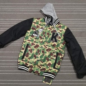 K&ouml;nnte beinhalten: Eine gr&uuml;ne, schwarze und wei&szlig;e Tarnjacke mit Kapuze und einem gro&szlig;en schwarzen Buchstaben "K" auf der Vorderseite. Die Jacke hat einen Rei&szlig;verschluss und eine Kapuze mit Kordelzug.