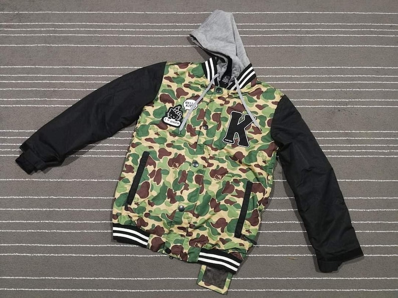 K&ouml;nnte beinhalten: Eine gr&uuml;ne, schwarze und wei&szlig;e Tarnjacke mit Kapuze und einem gro&szlig;en schwarzen Buchstaben "K" auf der Vorderseite. Die Jacke hat einen Rei&szlig;verschluss und eine Kapuze mit Kordelzug.