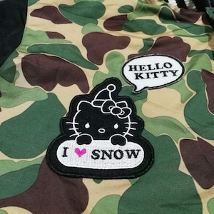 K&ouml;nnte beinhalten: Eine gr&uuml;n-braune Tarnjacke mit schwarzen &Auml;rmeln. Die Jacke hat einen Hello Kitty-Aufn&auml;her mit dem Text "HELLO KITTY" und einen Aufn&auml;her mit einer Hello Kitty-Illustration und dem Text "I &hearts; SNOW".