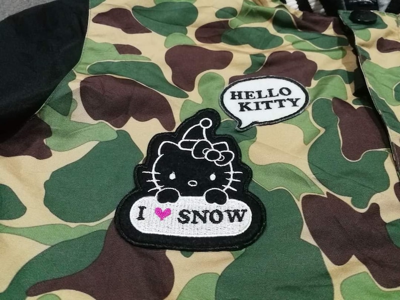 K&ouml;nnte beinhalten: Eine gr&uuml;n-braune Tarnjacke mit schwarzen &Auml;rmeln. Die Jacke hat einen Hello Kitty-Aufn&auml;her mit dem Text "HELLO KITTY" und einen Aufn&auml;her mit einer Hello Kitty-Illustration und dem Text "I &hearts; SNOW".