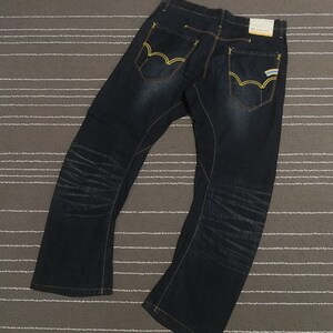 Può includere: Jeans in denim blu scuro con cuciture gialle e una piccola etichetta blu e bianca sulla tasca posteriore.