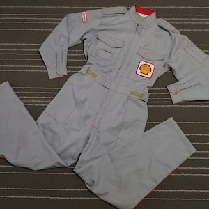 Pode incluir: Um macacão azul claro com guarnições vermelhas e um remendo com o logotipo da Shell. O macacão tem um fecho de correr e dois bolsos no peito. O logotipo da Shell é uma concha amarela e vermelha sobre um fundo branco.