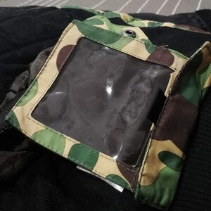 K&ouml;nnte beinhalten: Eine schwarze und gr&uuml;ne Tarnjacke mit einer durchsichtigen Plastiktasche auf der linken Seite. Die Tasche hat ein braunes, beiges und gr&uuml;nes Tarnmuster.