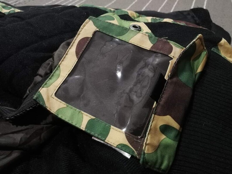 K&ouml;nnte beinhalten: Eine schwarze und gr&uuml;ne Tarnjacke mit einer durchsichtigen Plastiktasche auf der linken Seite. Die Tasche hat ein braunes, beiges und gr&uuml;nes Tarnmuster.